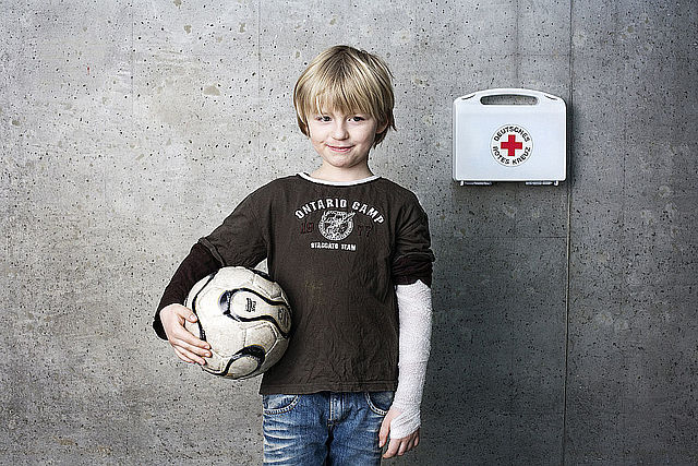 Junge mit verbundenem Arm hält einen Ball. Neben ihm ein Verbandskasten mit dem Logo des DRK