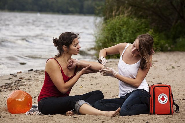 Szene am Strand: eine Frau verarztet die Wunde, die sich ihre Freundin am Ellbogen zugezogen hat. Vor ihr liegt ein Verbandset mit dem Logo des DRK.