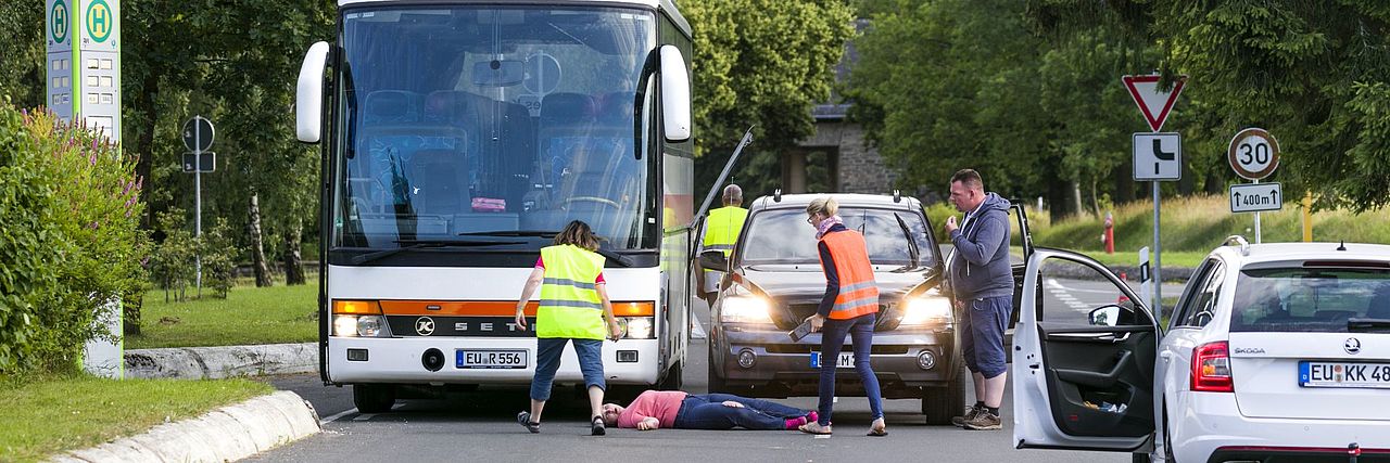 Erste Hilfe bei einem Verkehrsunfall, Fußgängerin liegt am Boden, nachdem sie die Straße vor einem Bus überqueren wollte. Ersthelfer sind vor Ort.