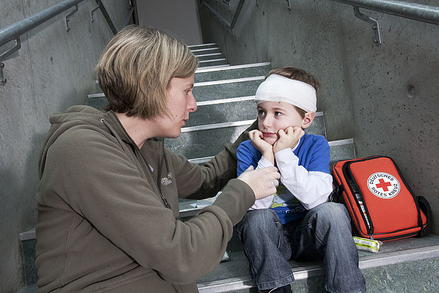 Mutter mit ihrem verletzten Sohn, er trägt einen Verband am Kopf. Neben dem Kind ein Verbandset mit dem Logo des DRK
